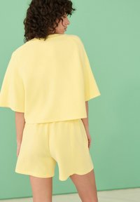 Haut jaune à manches courtes et short assorti, fabriqués en tissu doux avec une coupe décontractée. Fond en vert pastel. Détail en ourlet festonné.