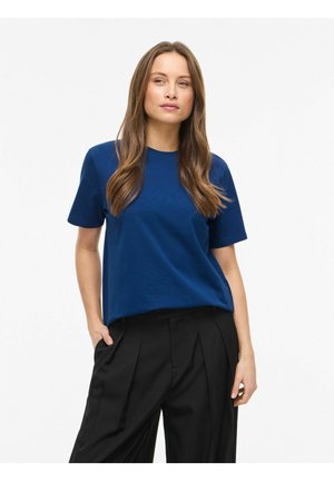 Femme aux longs cheveux bruns portant une chemise bleue à manches courtes et un pantalon noir plissé, debout avec une main dans la poche sur fond blanc.