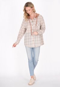Grijze en beige geruite blouse met capuchon, knoopsluiting aan de voorkant, lange mouwen en een losse fit, gecombineerd met lichtblauwe skinny jeans en beige sneakers.