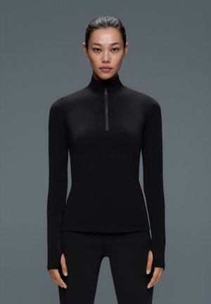 Une femme est de face, vêtue d’un haut de sport noir à manches longues avec un col montant zippé et d’un legging noir, sur un fond gris.