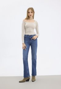 Beige Langarmoberteil mit schulterfreiem Design, kombiniert mit blauen geraden Jeans; olivgrüne Knöchelboots mit spitzen Zehen; minimalistischer Hintergrund.