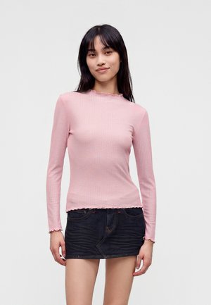 Kvinde med langt sort hår iført en tætsiddende lyserød langærmet top og et kort mørkt denimskørt, der står mod en ensfarvet baggrund.