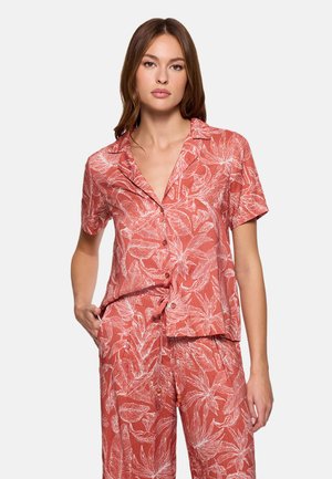 Mujer con camiseta de manga corta y pantalones de color coral con estampado de hojas tropicales blancas, de pie con una mano en el bolsillo sobre un fondo liso.