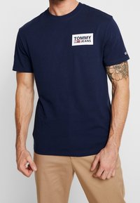 Navy blue cotton t-shirt with short sleeves, featuring a white rectangular logo with "TOMMY JEANS" text and red accent stripes on front.