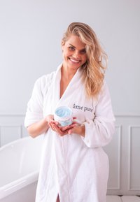 White terry cloth robe with "âme pure" emblem, holding a round, white container with a light blue cream and aqua label, set against a light background.