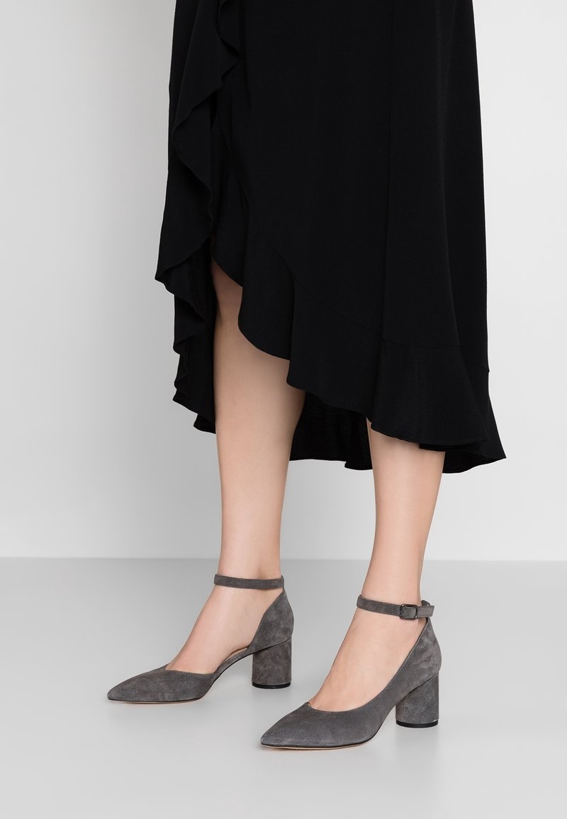 Pieds chaussés de chaussures à talon bloc en suède gris avec brides à la cheville, partiellement couverts par une jupe midi noire à volants.