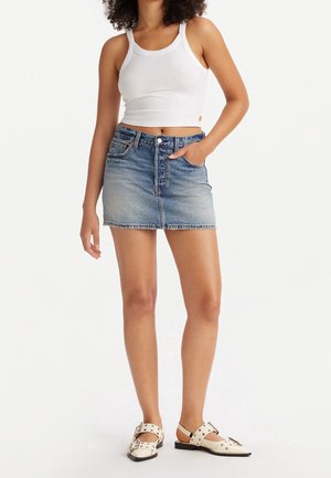 Femme portant un crop top blanc sans manches, une mini-jupe en jean bleu délavé et des ballerines crème cloutées, debout sur un fond blanc.