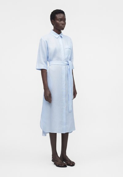 Γυναίκα που φοράει ανοιχτό μπλε πουκάμισο-φόρεμα με ζώνη, στέκεται όρθια μπροστά σε απλό λευκό φόντο.