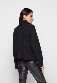Schwarzer, gerippter Strickpullover mit hohem Kragen, verkürztem Schnitt und langen Ärmeln, kombiniert mit glänzenden schwarzen Lederhosen.