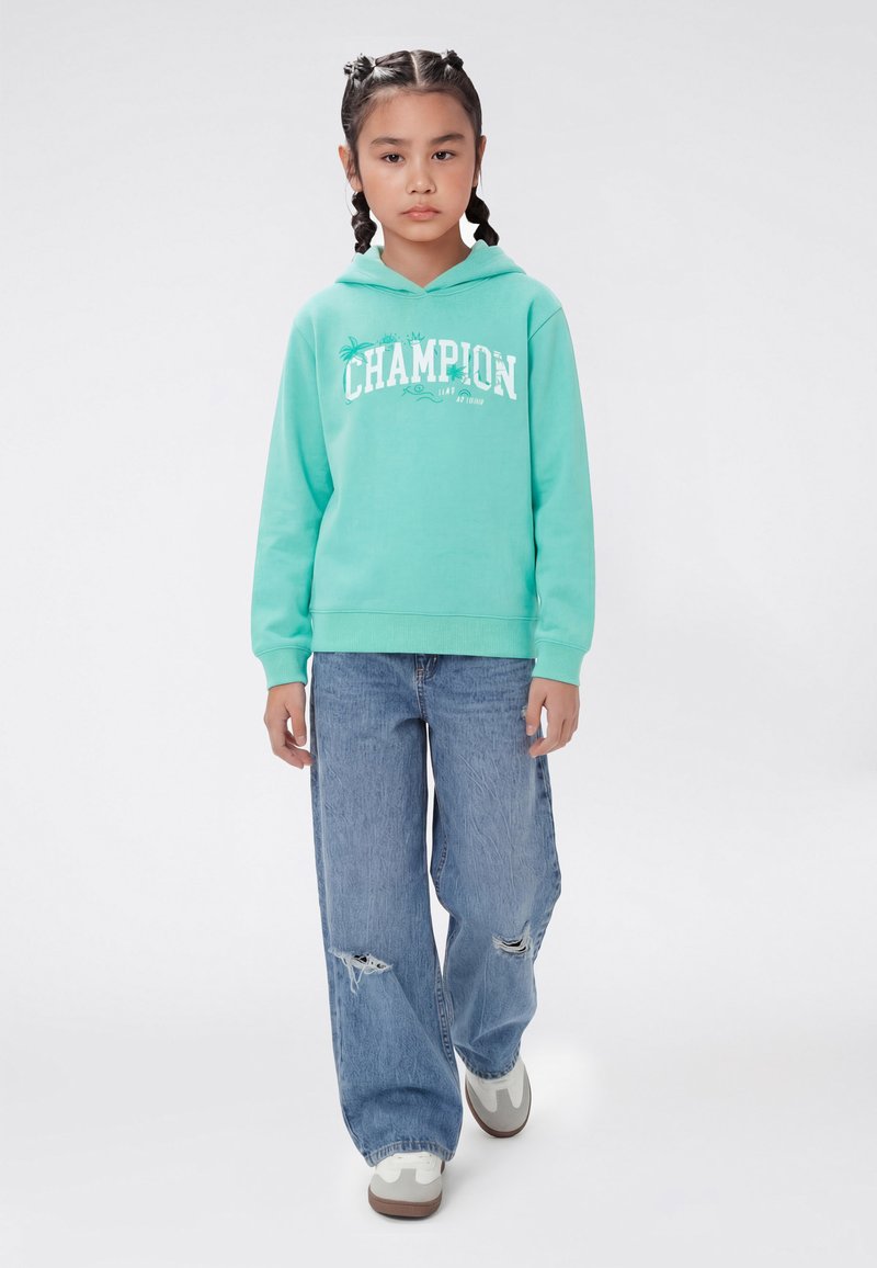 Fille aux cheveux tressés portant un sweat à capuche turquoise de la marque Champion, un jean à jambes larges déchiré et des baskets blanches, debout devant un fond blanc.