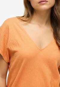 Pomarańczowa bluzka z dekoltem w serek, wykonana z teksturowanej, marszczonej tkaniny. Krótkie rękawy i luźny krój, eksponujące subtelne detale projektu.