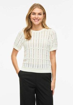 Mlada žena smiješi se, nosi bijelu pamučnu majicu kratkih rukava s teksturiranim uzorkom i crne hlače, stoji s jednom rukom u džepu.