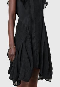 Robe noire avec des panneaux texturés et un détail lisse à l'avant portée par une personne à la peau foncée, montrant le bras et la main avec un bracelet délicat.