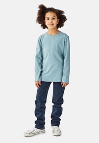 Lyseblå langærmet t-shirt lavet af bomuld, kombineret med mørkeblå jeans og hvide sneakers, stående foran en neutral baggrund.