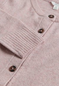 Lichtroze gebreide cardigan met een ronde halslijn, gestreepte ribkappen en donkerbruine knopen. Zacht, donzig materiaal met subtiele variaties.