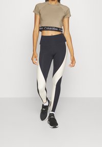 Brązowy krótki t-shirt z czarną opaską Calvin Klein, czarno-białe legginsy z wysokim stanem i geometrycznym wzorem, noszone z czarnymi butami sportowymi.