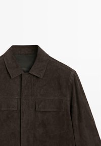 Giacca in camoscio marrone con colletto, due tasche sul petto e chiusura frontale con bottoni. Tessuto liscio con dettagli di cuciture visibili.