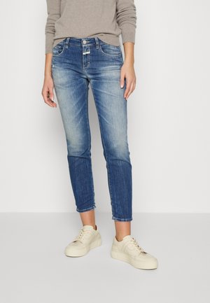 Personne portant un jean bleu délavé, un pull beige et des baskets couleur crème à lacets, debout sur un fond simple de sol et de mur clairs.