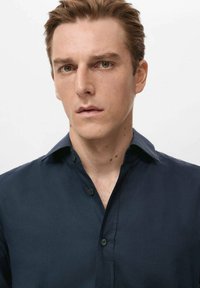 Camisa azul marino abotonada con una textura suave, que presenta un cuello clásico y botones negros, llevada por un modelo masculino con expresión neutral.