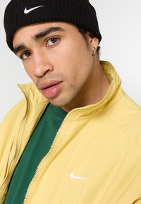 Gul zip-up jakke med et hvidt Nike-logo, båret over en grøn t-shirt, parret med en sort strikbeanie. Glat tekstur og afslappet pasform.