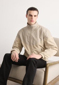 Casaco bege com fecho zip e painel superior estampado, punhos elásticos e bolsos laterais, combinado com calças pretas. Textura suave, design moderno.