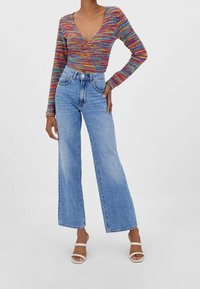 Meerkleurige, langemouwige wikkeltop met diepe V-hals, gecombineerd met high-waisted, wijde lichtblauwe jeans en witte sandalen met bandjes.