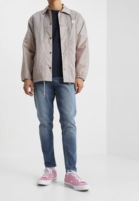 Chaqueta ligera de color gris claro con cierre de botones a presión y puños elásticos, usada sobre una camisa azul marino oscura. Combinada con jeans azules y zapatillas rosas.