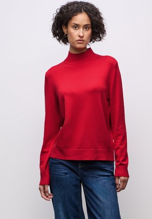 Czerwony sweter z dzianiny o wysokim dekolcie i długich rękawach; gładka faktura. Połączony z niebieskimi dżinsami. Prosty i nowoczesny design.