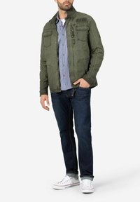 Olivgrüne Jacke mit Reißverschluss, vier Taschen und Stehkragen. Getragen über einem blau gestreiften Hemd, kombiniert mit dunklen Jeans und weißen Sneakers.