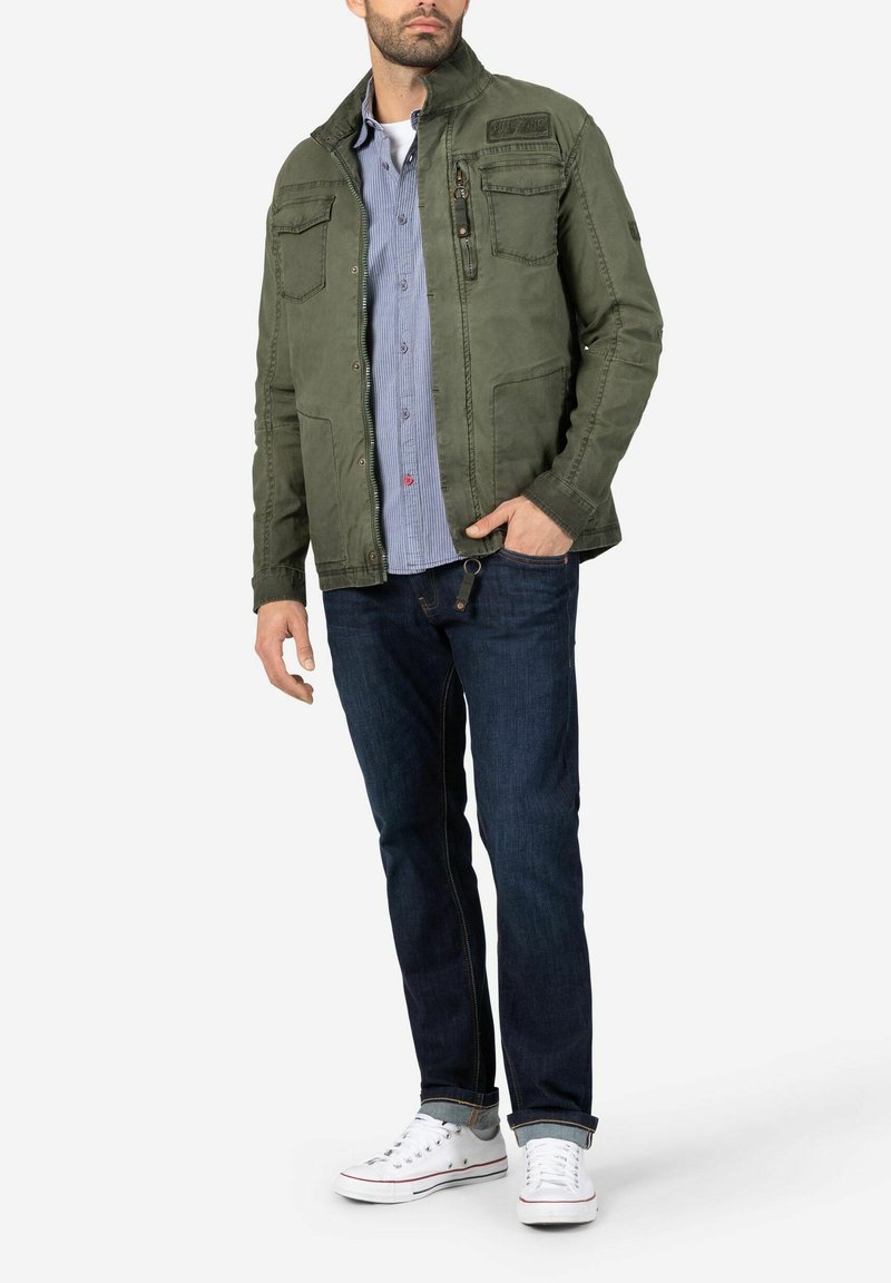 Olivgrüne Jacke mit Reißverschluss, vier Taschen und Stehkragen. Getragen über einem blau gestreiften Hemd, kombiniert mit dunklen Jeans und weißen Sneakers.