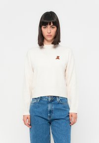 Pulover ribbed de culoare crem, cu guler rotund și un mic broderie cu un ursuleț de pluș maro pe piept, asortat cu blugi albaștri cu talie înaltă.