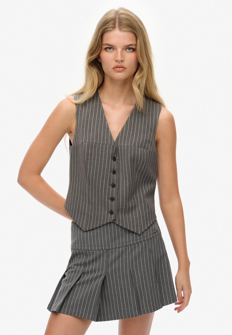 Szary kamizelka w prążki z głębokim dekoltem w kształcie litery V, z pięcioma ciemnymi guzikami i dopasowanym krojem, w komplecie ze spódnicą plisowaną o wysokim stanie, pasującą kolorystycznie.