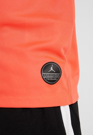 Nahaufnahme eines korallenfarbenen sportlichen Shirt-Saums mit einem schwarzen runden Aufnäher mit der Aufschrift "Engineered for Championship Athletes" über schwarz-weißen Shorts.