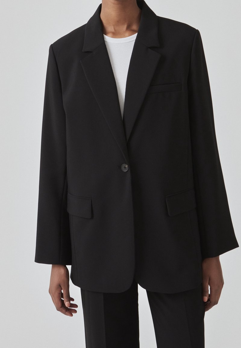 Personne portant un blazer noir à un bouton sur une chemise blanche côtelée, montrée des épaules jusqu'à mi-cuisse sur un fond uni.