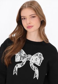 Fekete pulóver, közepén fehér csipkés masni mintával. Puha anyagból, kerek nyakkivágással és laza szabással.