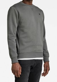 Šedá mikina s kulatým výstřihem, žebrovanými lemy na rukávech a spodním okraji. Na hrudi má malý vyšívaný logo a hladkou texturu.