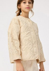 Quiltad beige jacka med ett broderat hästmönster, rund hals och manschetterade ärmar för en texturerad finish och strukturerad design.