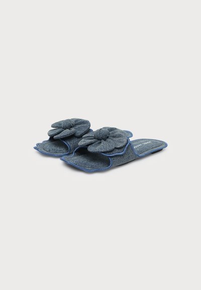 Paire de pantoufles en denim bleu à enfiler avec de grandes fleurs en tissu sur le dessus, bords arrondis et liseré bleu, présentées sur fond blanc.