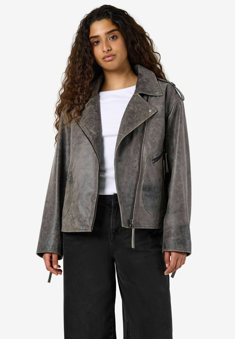 Jeune femme aux longs cheveux bouclés portant une veste de motard en suède gris par-dessus une chemise blanche et un pantalon noir sur un fond uni.
