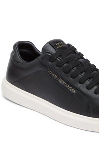 Čierna kožená teniska s plochými šnúrkami, polstrovaným golierom a bielou gumovou podrážkou. Obsahuje vyrazené značkovanie „TOMMY HILFIGER“ v zlatej farbe.