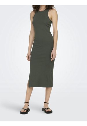 Frau trägt ärmelloses, dunkelgrünes, geripptes Midikleid und schwarze Plateau-Sandalen und steht vor einem einfarbigen, hellen Hintergrund