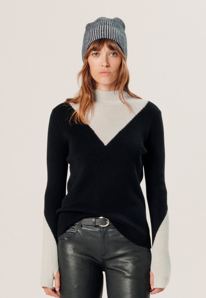 Zwart en grijs blokgestructureerd sweater met een hoge hals, met lange mouwen met contrasterende kleuraccenten. Model draagt een gestreepte beanie.