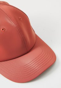 Une casquette de couleur corail avec une couronne structurée et une visière cousue repose sur une surface blanche, mettant en avant sa texture lisse et ses détails.