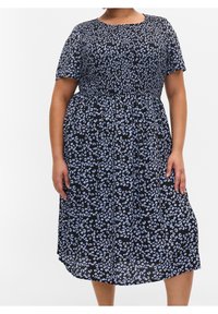 Osoba w rozmiarze plus nosi czarną sukienkę do kolan z niebieskim kwiatowym wzorem i krótkimi rękawami, stojąc na tle białym.