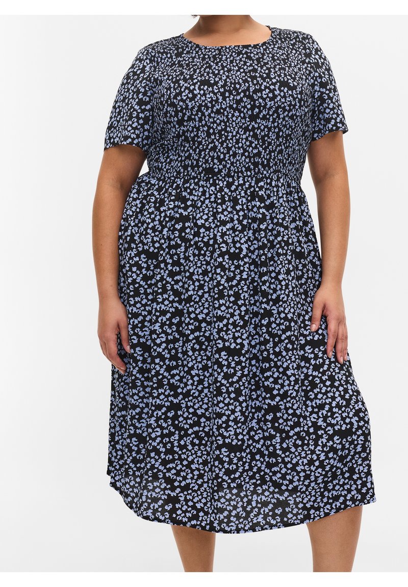 Osoba w rozmiarze plus nosi czarną sukienkę do kolan z niebieskim kwiatowym wzorem i krótkimi rękawami, stojąc na tle białym.