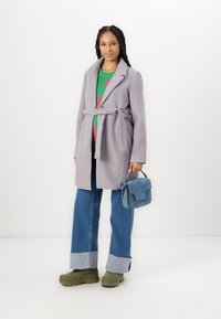 Grå belted frakk over en grønn genser med rosa design, sammen med brede denimbukser og grønne chunky sko. Liten blå veske som aksent.