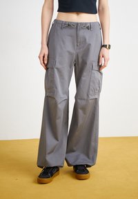 Šedé cargo kalhoty s širokými nohavicemi, stahovací pas, více postranními kapsami a hladkým bavlněným povrchem. Nosí se s černými botami na žlutém pozadí.
