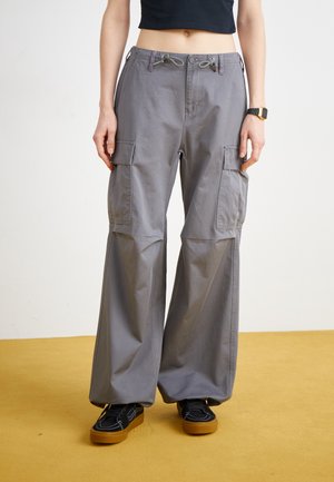 Szare spodnie cargo o szerokich nogawkach, z wiązanym pasem, licznymi kieszeniami bocznymi i gładką bawełnianą teksturą. Noszone z czarnymi butami na żółtym tle.