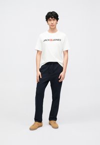 Camiseta blanca de manga corta con logo negro "JACK & JONES", combinada con pantalones deportivos azul marino y zapatillas de color marrón claro sobre un fondo liso y claro.