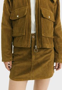 Frau trägt eine braune Cord-Jacke und einen passenden Rock mit Reißverschluss und Knopfdetails, kombiniert mit einem weißen, gerippten Oberteil.